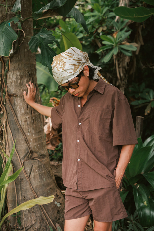 A person stands barefoot outdoors, leaning against a tree in a tropical setting. They are dressed in a brown short-sleeved button-up shirt and matching shorts. A patterned scarf is tied around their head, and they are wearing dark sunglasses. The background is filled with dense green foliage and tropical plants, creating a relaxed, nature-inspired atmosphere.