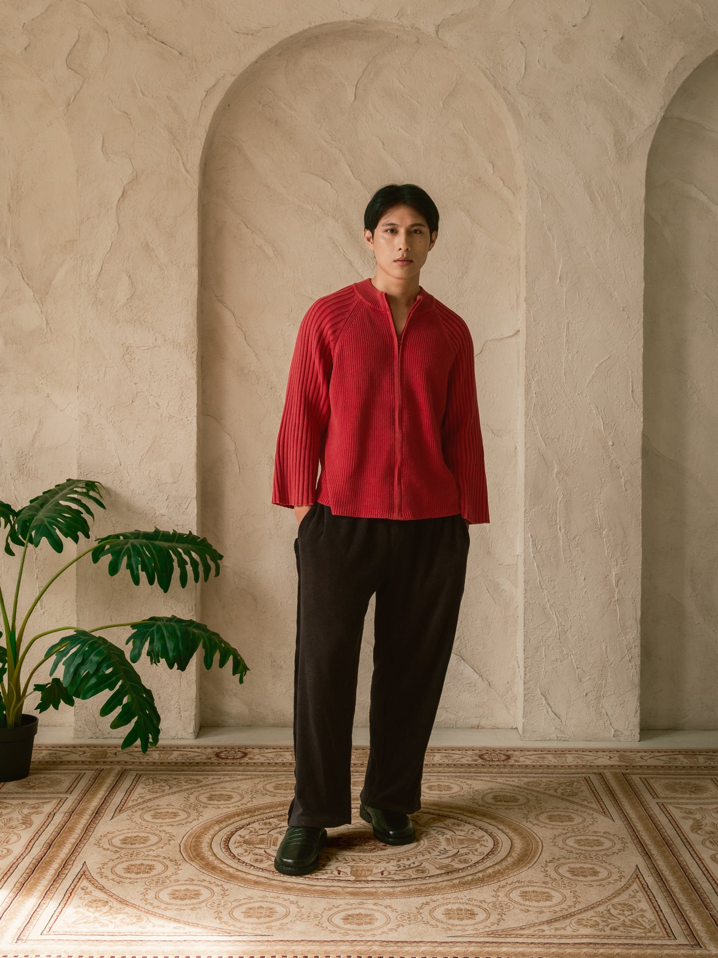 Person wearing a red shirt and black pants standing in a room with decorative arches and a plant.
