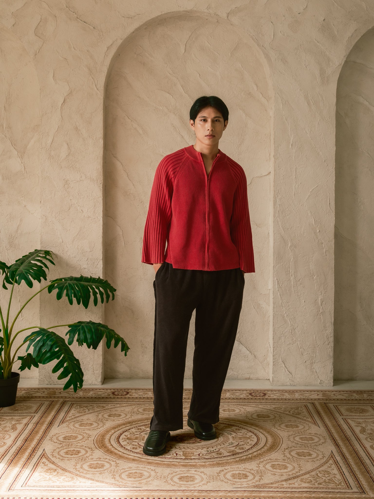 Person wearing a red shirt and black pants standing in a room with decorative arches and a plant.
