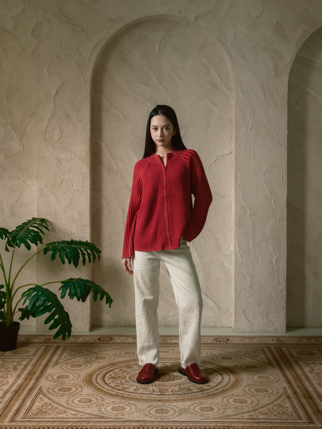 Woman in a red blouse and white pants standing in a room with decorative arches and a plant.