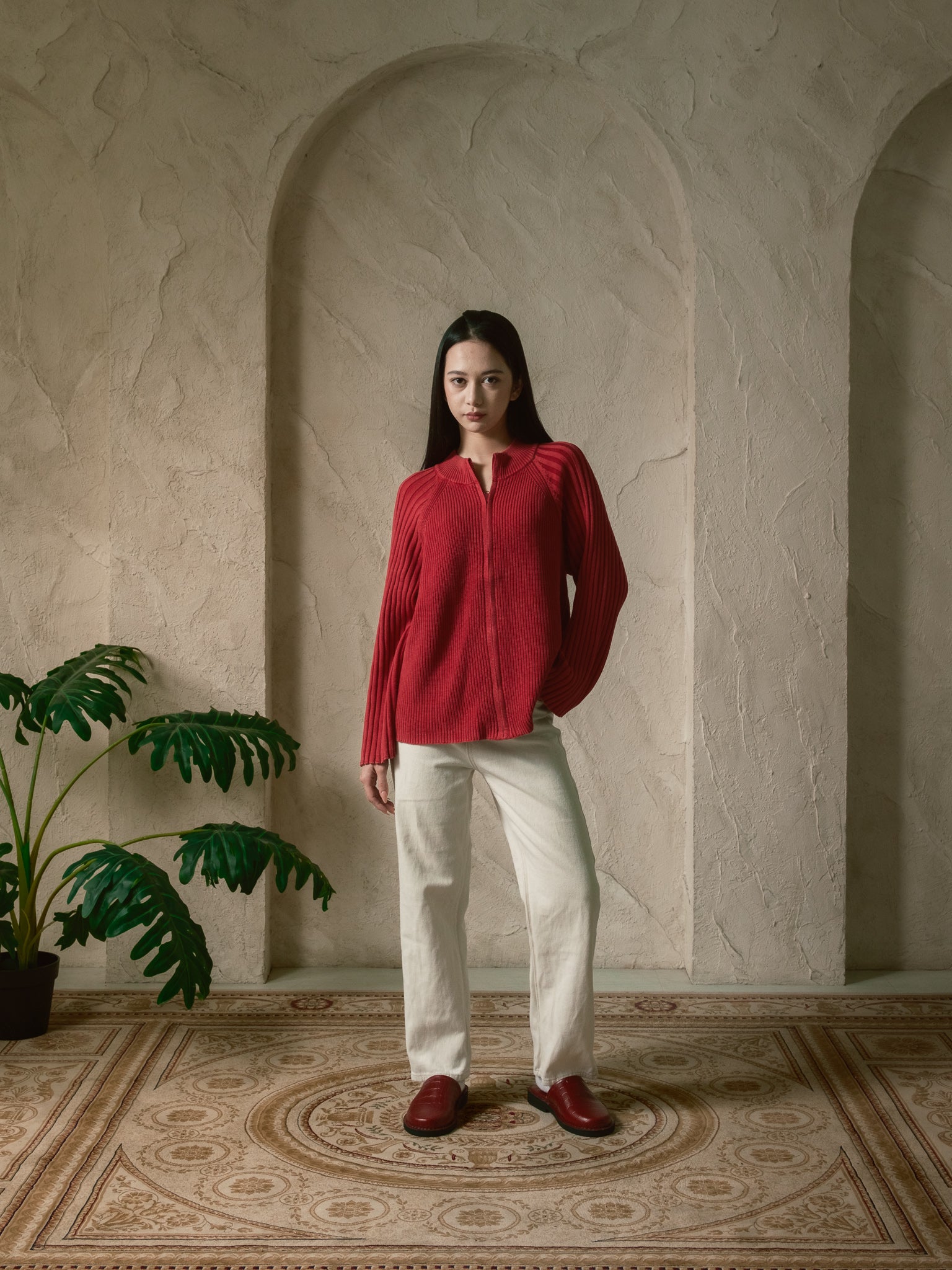 Woman in a red blouse and white pants standing in a room with decorative arches and a plant.