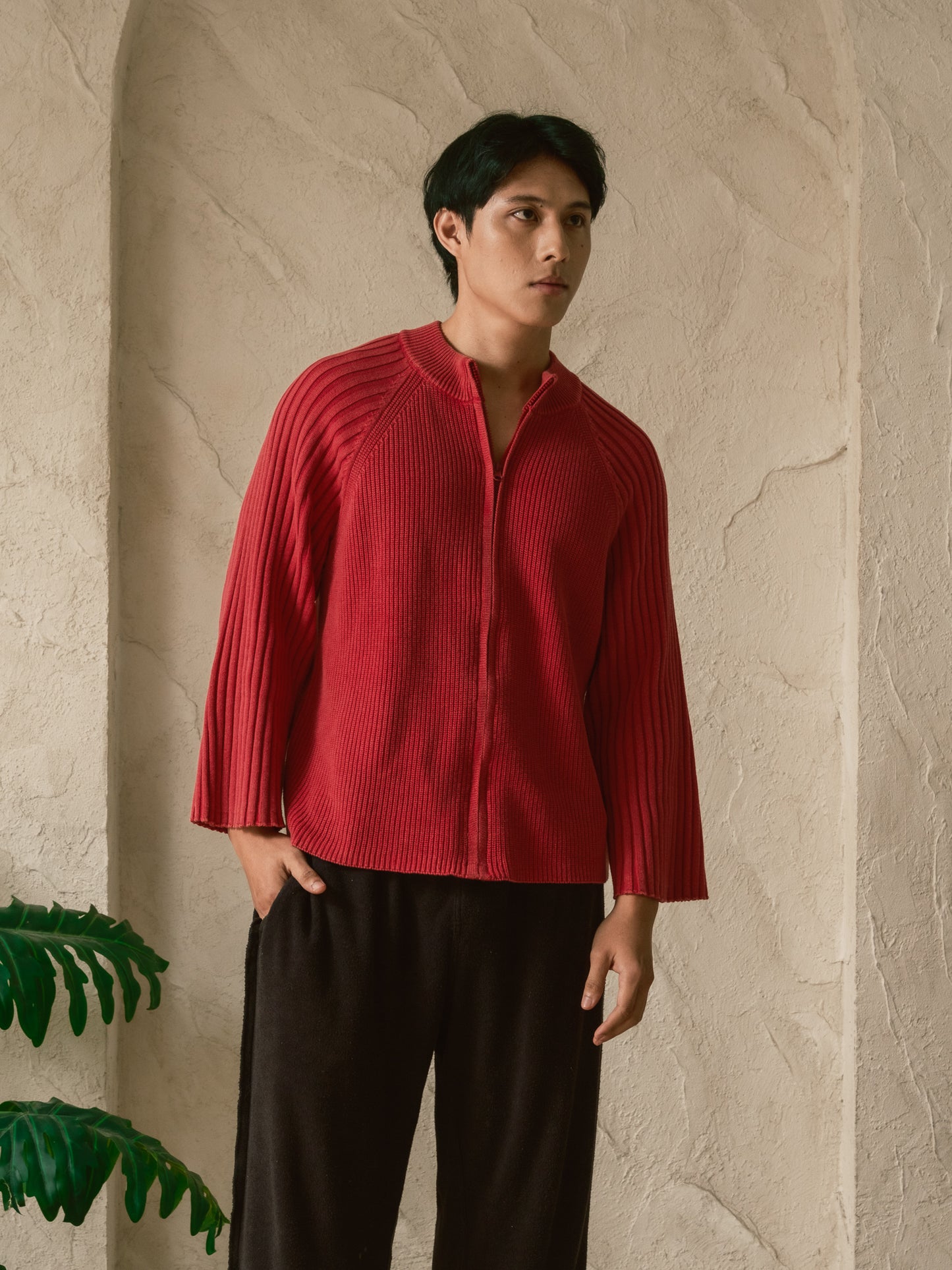Person wearing a red ribbed sweater and black pants against a beige wall.