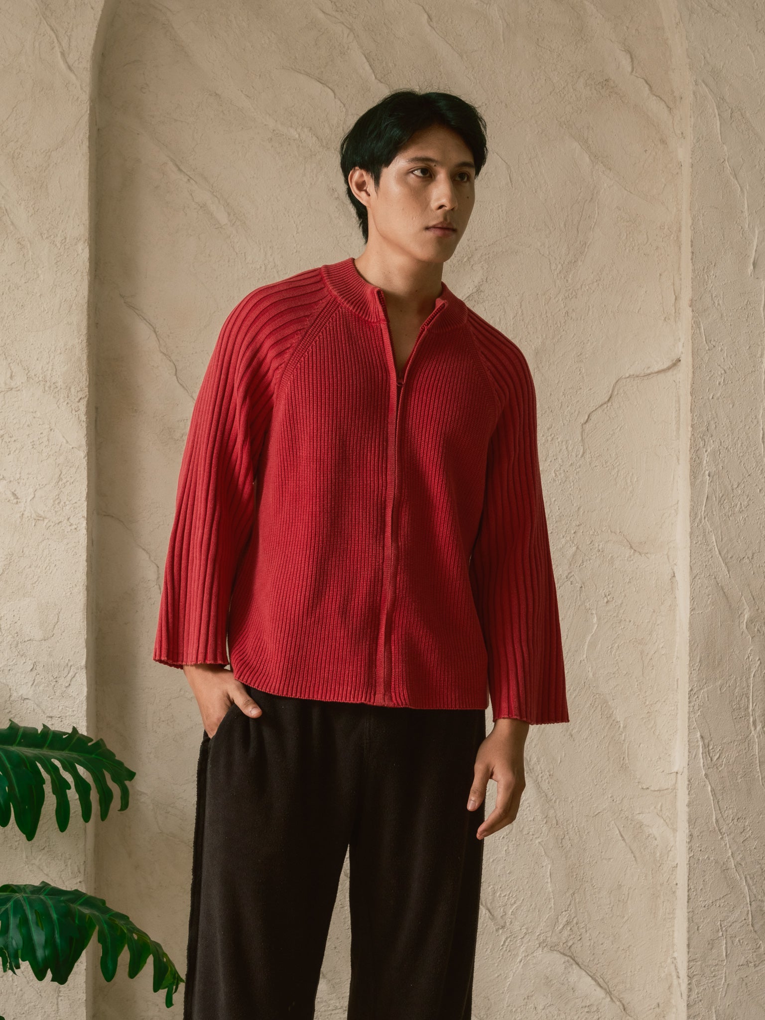 Person wearing a red ribbed sweater and black pants against a beige wall.