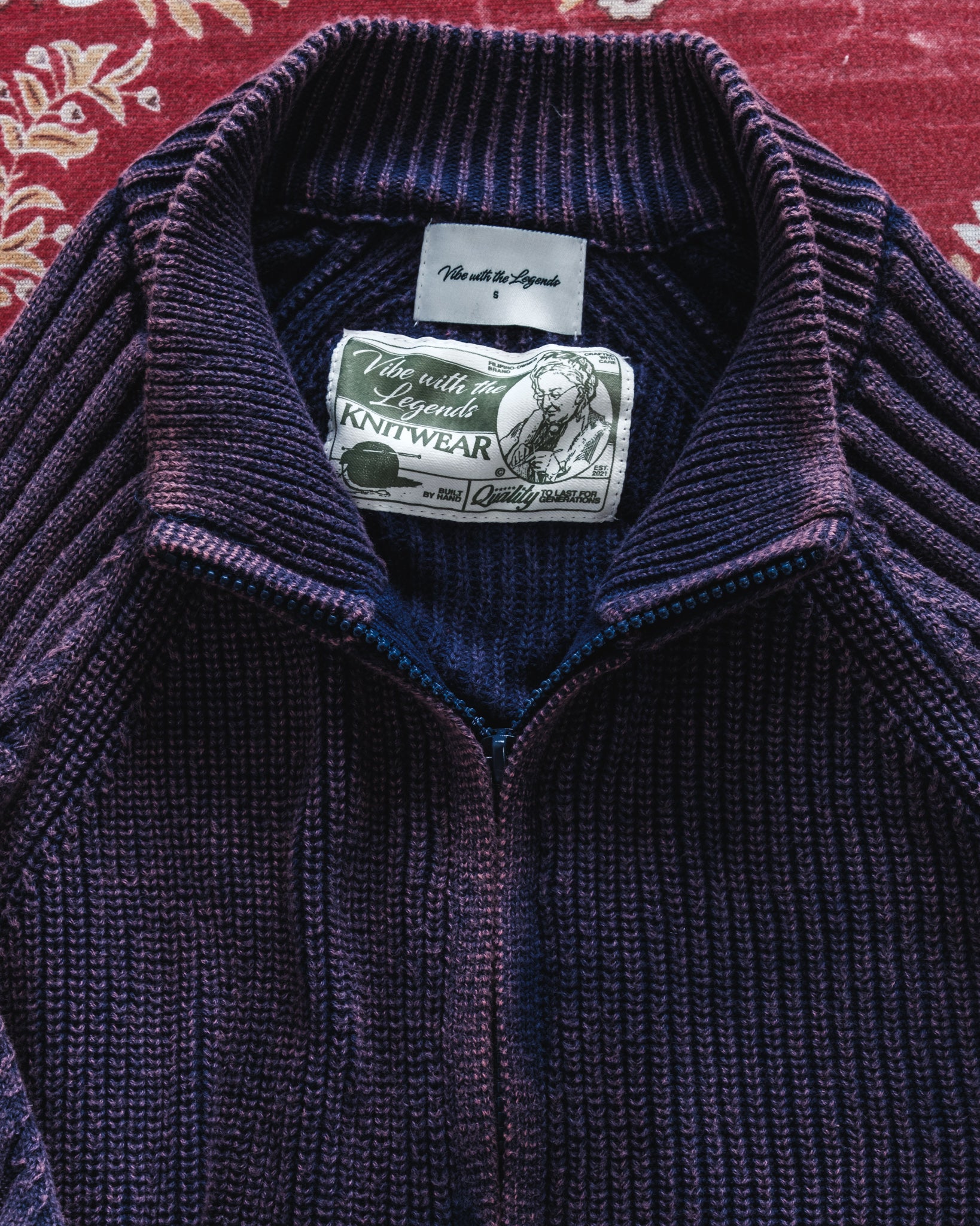 Close-up of a purple knit sweater with a brand label on a red patterned background