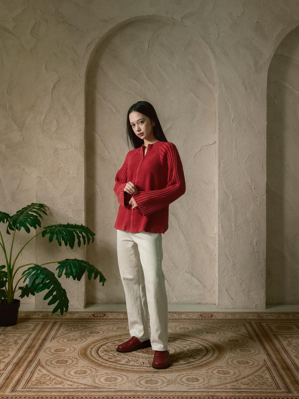 Woman in a red blouse and white pants standing in a room with a plant and patterned floor.