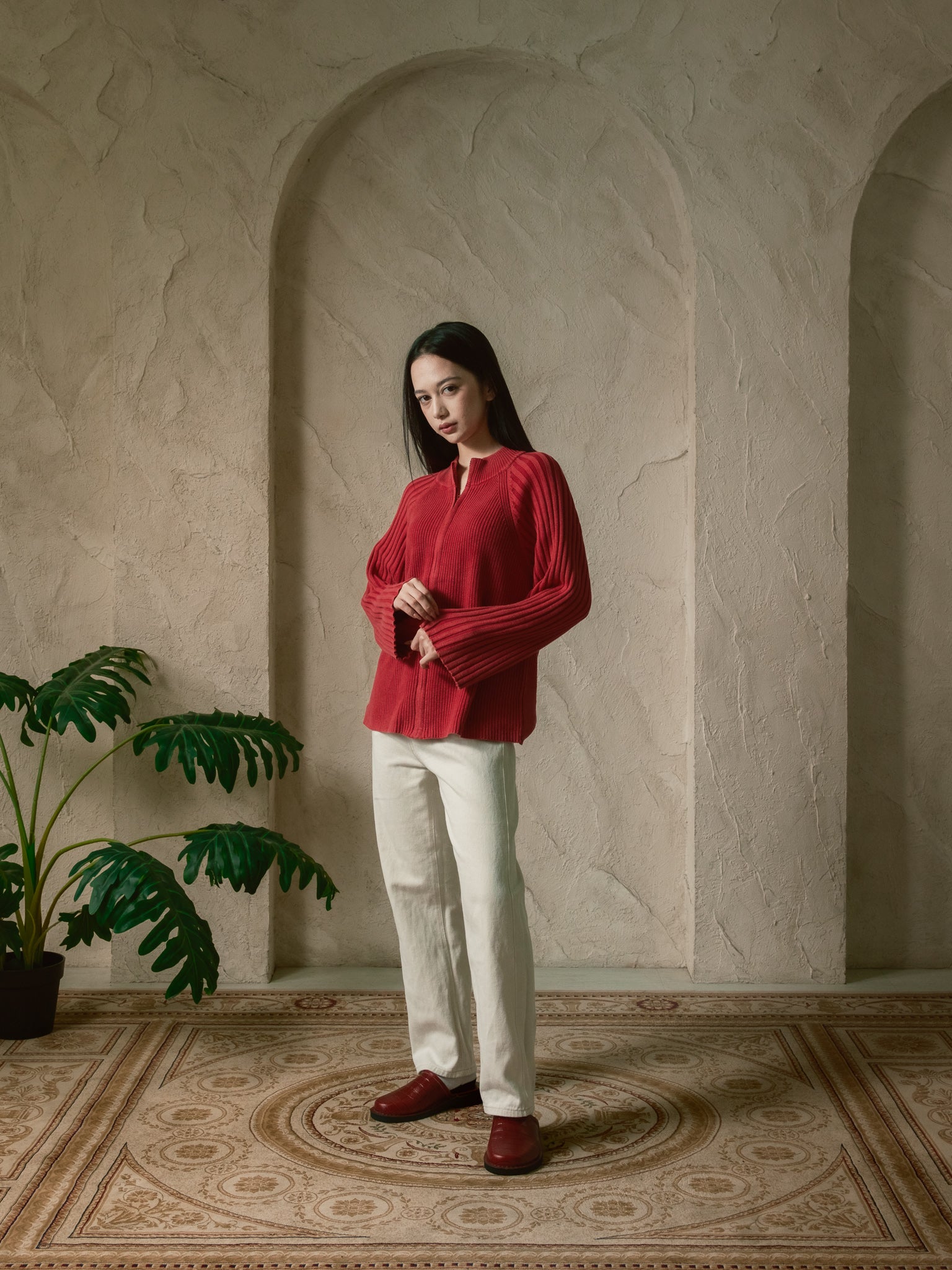 Woman in a red blouse and white pants standing in a room with a plant and patterned floor.