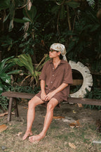  A person sits barefoot on a wooden bench outdoors, surrounded by lush tropical foliage. They are wearing a loose brown short-sleeved button-up shirt and matching shorts, along with sunglasses and a patterned scarf wrapped around their head. A large white circular object, possibly a repurposed tire, rests against the greenery in the background. The setting has a relaxed, natural vibe with fallen leaves scattered on the ground.