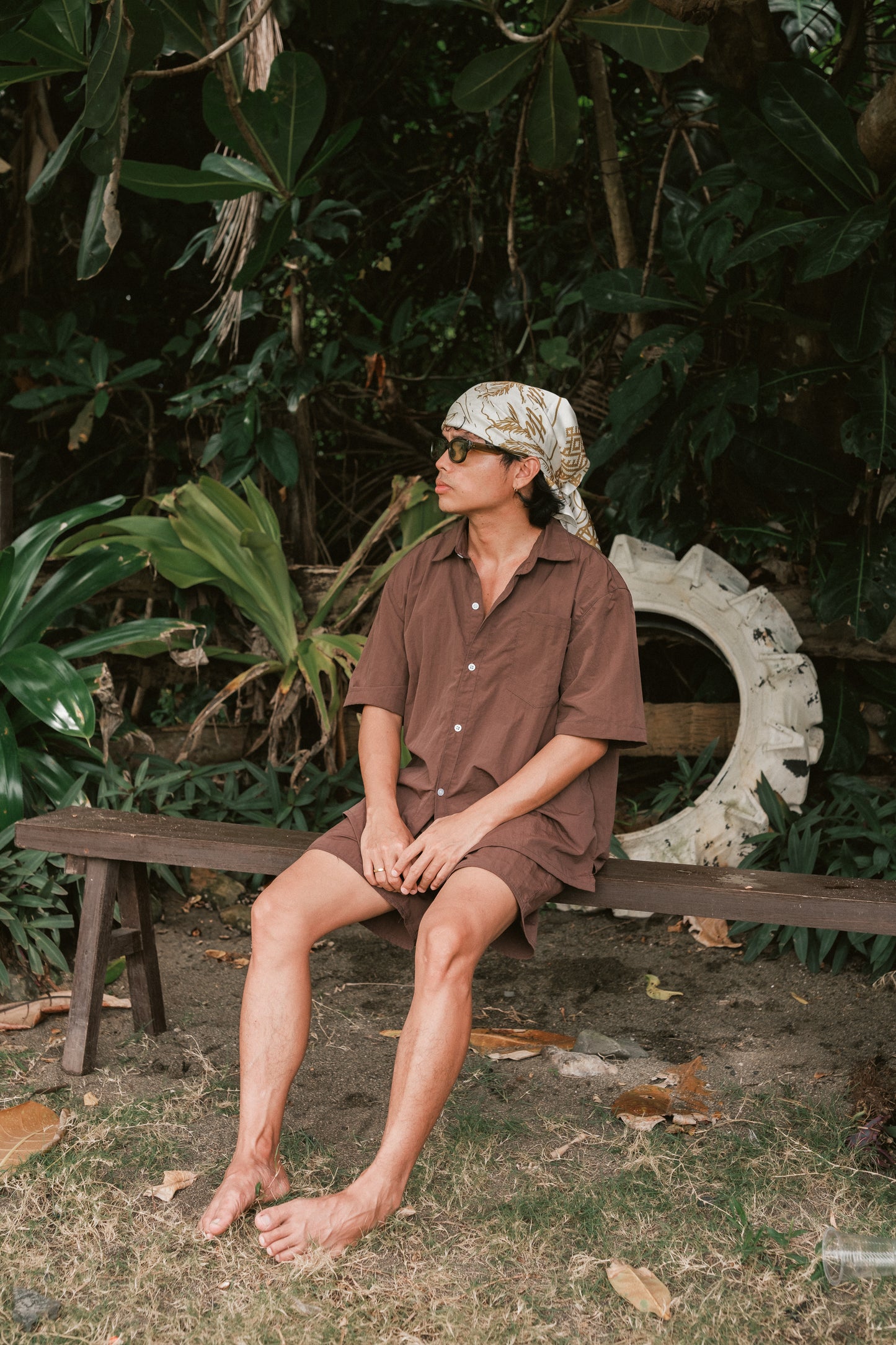 A person sits barefoot on a wooden bench outdoors, surrounded by lush tropical foliage. They are wearing a loose brown short-sleeved button-up shirt and matching shorts, along with sunglasses and a patterned scarf wrapped around their head. A large white circular object, possibly a repurposed tire, rests against the greenery in the background. The setting has a relaxed, natural vibe with fallen leaves scattered on the ground.
